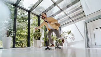Mann in Anzug fährt auf einem Skateboard in einem modernen Raum mit großen Fenstern und Pflanzen.