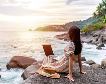 Eine Frau sitzt auf einem Felsen am Meer, mit einem Laptop auf den Knien und einem Hut neben sich, während die Sonne untergeht.