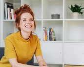 Eine lächelnde Frau mit roten Haaren sitzt in einem modernen Raum, umgeben von Regalen mit Büchern und Pflanzen. Sie trägt ein gelbes Oberteil und wirkt entspannt.