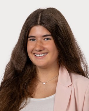 Junge Frau mit langen, lockigen Haaren, die in einem hellen Blazer und einem weißen Oberteil lächelt. Neutraler Hintergrund.