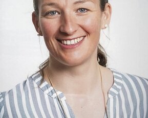 Eine lächelnde Frau mit blonden Haaren trägt ein gestreiftes Hemd. Sie hat Ohrringe und schaut direkt in die Kamera.
