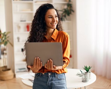 Eine Frau mit lockigem Haar hält einen Laptop in den Händen und lächelt. Sie trägt ein orangefarbenes Hemd und steht in einem hellen Raum mit Pflanzen und einem Tisch.