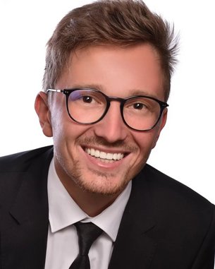 Ein junger Mann mit Brille und einem Anzug lächelt freundlich in die Kamera. Sein Haar ist kurz und leicht zerzaust. Der Hintergrund ist einfarbig und neutral.