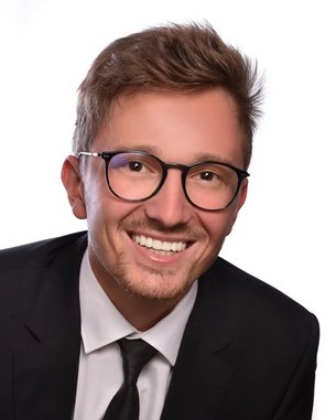 Ein junger Mann mit Brille und einem Anzug lächelt freundlich in die Kamera. Sein Haar ist kurz und leicht zerzaust. Der Hintergrund ist einfarbig und neutral.