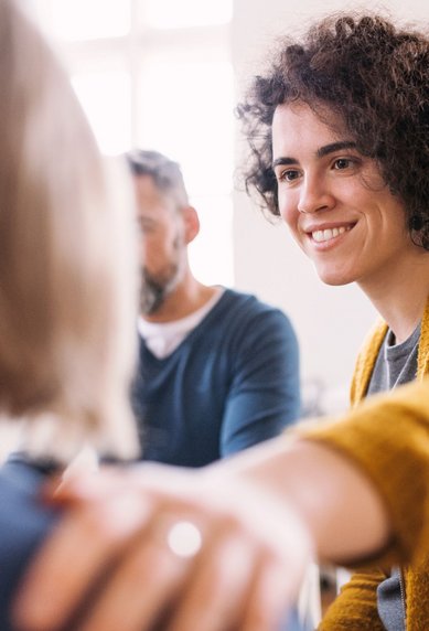 Eine lächelnde Frau mit lockigem Haar trägt einen gelben Pullover und legt ihre Hand auf die Schulter einer anderen Person. Im Hintergrund sind zwei weitere Personen zu sehen, die an einem Gespräch teilnehmen.