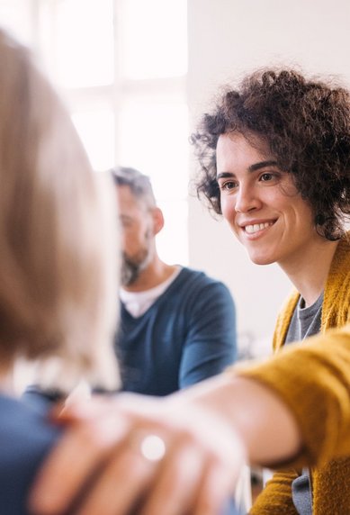 Eine lächelnde Frau mit lockigem Haar trägt einen gelben Pullover und legt ihre Hand auf die Schulter einer anderen Person. Im Hintergrund sind zwei weitere Personen zu sehen, die an einem Gespräch teilnehmen.