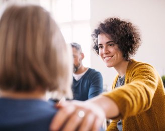 Positives Denken in der zwischenmenschlichen Kommunikation Eine lächelnde Frau mit lockigem Haar trägt einen gelben Pullover und legt ihre Hand auf die Schulter einer anderen Person. Im Hintergrund sind zwei weitere Personen zu sehen, die an einem Gespräch teilnehmen.