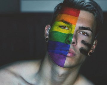 Ein junger Mann mit einem Gesicht, das in Regenbogenfarben bemalt ist, steht im Vordergrund. Er hat kurze, graue Haare und trägt schwarze Ohrstecker. Ein Teil seines Gesichts ist mit schwarzer Farbe markiert.