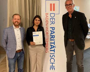 Drei Personen stehen vor einem Banner mit der Aufschrift "DER PARITÄTISCHE BADEN-WÜRTTEMBERG". Eine Person hält ein Zertifikat in der Hand, während die anderen beiden neben ihr stehen.