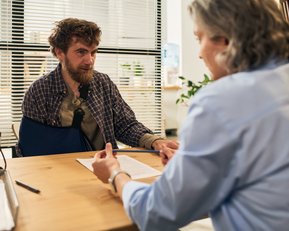 Ein Mann mit einem Verband am Arm sitzt an einem Tisch und spricht mit einer Person, die ihm gegenüber sitzt.