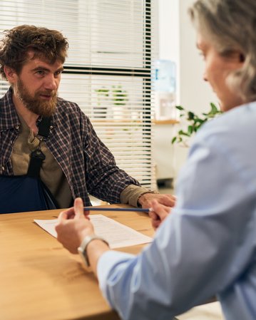 Ein Mann mit einem Verband am Arm sitzt an einem Tisch und spricht mit einer Person, die ihm gegenüber sitzt.