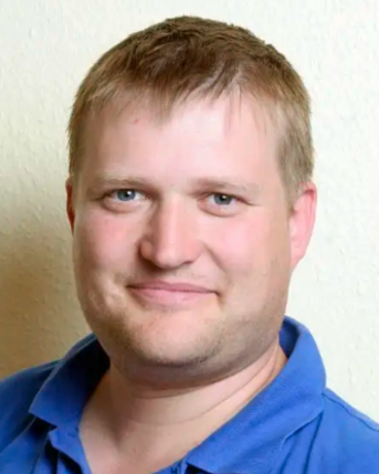 Ein Mann mit kurzen, blonden Haaren und einem freundlichen Gesichtsausdruck trägt ein blaues Hemd. Der Hintergrund ist hell und neutral.