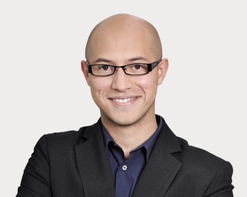 Mann mit Glatze und Brille, trägt einen schwarzen Anzug und ein dunkles Hemd, lächelt freundlich und steht mit verschränkten Armen.