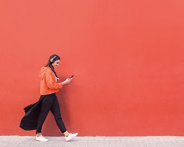 Studiengänge ohne NC Eine Person mit Kopfhörern trägt einen orangefarbenen Hoodie und schwarze Hosen, während sie an einer roten Wand entlanggeht und ein Smartphone in der Hand hält.