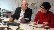Zwei Personen sitzen an einem Tisch, der mit Notizen, einem Telefon und Wasserflaschen bedeckt ist. Sie diskutieren und lächeln, während sie Notizen machen und sich austauschen.