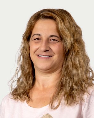 Eine Frau mit lockigem, braunem Haar lächelt freundlich in die Kamera. Sie trägt ein helles T-Shirt und hat einen neutralen Hintergrund.
