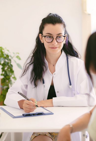 Eine Frau in einem weißen Kittel sitzt an einem Tisch und spricht mit einer Patientin, während sie Notizen macht.