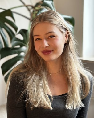 Eine junge Frau mit langen, blonden Haaren lächelt in die Kamera, während sie in einem hellen Raum vor einer Pflanze sitzt.