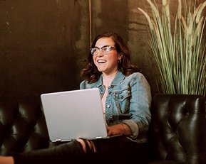 Eine Person mit Brille sitzt auf einer Ledercouch und arbeitet an einem Laptop. Im Hintergrund ist eine Pflanze sichtbar.
