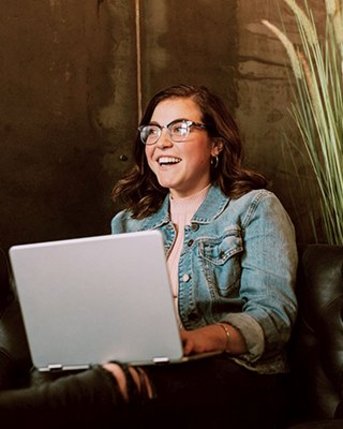 Eine Person mit Brille sitzt auf einer Ledercouch und arbeitet an einem Laptop. Im Hintergrund ist eine Pflanze sichtbar.