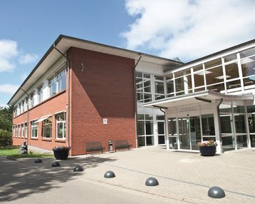 Moderne, zweigeschossige Gebäude mit roten Ziegeln und großen Fenstern, umgeben von Grünflächen und Sitzgelegenheiten im Freien.