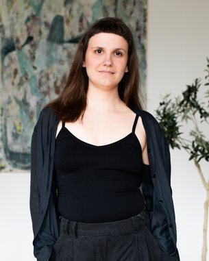 Eine Person mit langen, braunen Haaren trägt ein schwarzes Top und eine schwarze Jacke, steht vor einem abstrakten Kunstwerk und einer Pflanze.