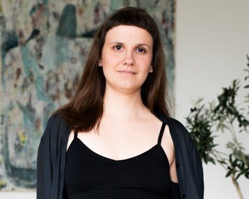 Eine Person mit langen, braunen Haaren trägt ein schwarzes Top und eine schwarze Jacke, steht vor einem abstrakten Kunstwerk und einer Pflanze.