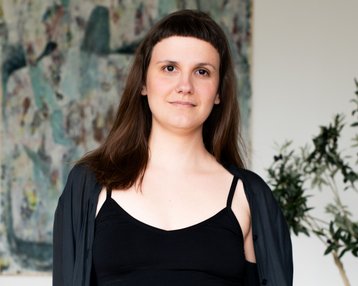 Eine Person mit langen, braunen Haaren trägt ein schwarzes Top und eine schwarze Jacke, steht vor einem abstrakten Kunstwerk und einer Pflanze.