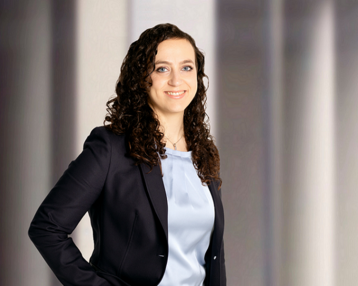 Kristin Rost, Studentin Psychologie (B.Sc.) | SRH Fernhochschule Eine lächelnde Frau mit lockigem, braunem Haar trägt einen schwarzen Blazer und ein hellblaues Oberteil. Sie steht mit einer Hand an der Hüfte vor einem neutralen Hintergrund.