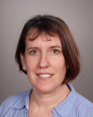 Eine Frau mit schulterlangen braunen Haaren und einem blauen Hemd lächelt in die Kamera. Der Hintergrund ist neutral und grau.