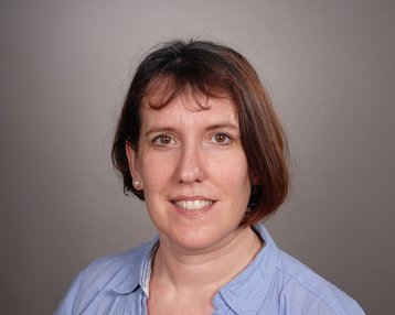 Eine Frau mit schulterlangen braunen Haaren und einem blauen Hemd lächelt in die Kamera. Der Hintergrund ist neutral und grau.