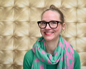 Eine Frau mit Brille und einem bunten Schal mit Herzen lächelt vor einem goldfarbenen, strukturierten Hintergrund.