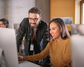 Ein Mann und eine Frau sitzen an einem Computer. Der Mann lächelt und schaut auf den Bildschirm, während die Frau konzentriert arbeitet. Beide scheinen in einem modernen Büro oder Lernumfeld zu sein.