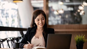 Eine Frau sitzt an einem Tisch, arbeitet an einem Laptop und macht Notizen in einem Notizbuch. Sie trägt einen schwarzen Blazer und lächelt, während sie auf den Bildschirm schaut.