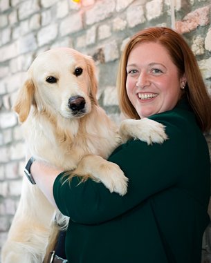 Eine Frau mit langen, roten Haaren hält einen Golden Retriever in ihren Armen. Sie lächelt und steht vor einer gemauerten Wand.