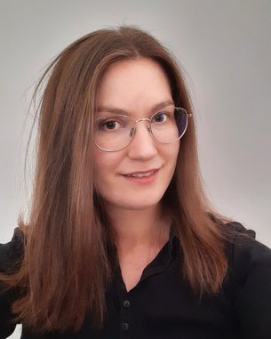 Eine Person mit langen, glatten Haaren und einer schwarzen Bluse trägt runde Brille und lächelt leicht in die Kamera. Der Hintergrund ist neutral und hell.