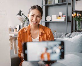 Eine Frau sitzt auf einem Sofa und spricht in ein Smartphone, während sie in einem modernen Raum mit Pflanzen und Bücherregalen umgeben ist. Im Hintergrund ist ein Flipchart sichtbar.