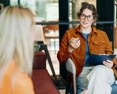 Berufe in der klinischen Psychologie ohne Approbation Eine Frau mit Brille und braunem Blazer sitzt auf einem Stuhl und spricht mit einer anderen Person, während sie ein Notizblatt hält.