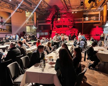 Ein geschäftliches Event mit zahlreichen Gästen, die an Tischen sitzen und essen, in einer großen Halle mit rotem Licht und industriellem Ambiente.
