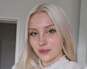 Eine Person mit langen, blonden Haaren trägt eine helle Jacke und schaut in die Kamera. Der Hintergrund zeigt einen neutralen Raum mit einer Tür.