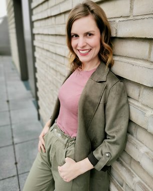Eine Frau mit lockigem, braunem Haar trägt ein rosa Oberteil und einen olivgrünen Blazer. Sie lehnt an einer hellen Ziegelwand und lächelt. Ihre Hände sind in den Hosentaschen.
