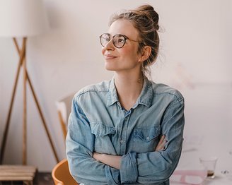 Junge Frau mit Brille und einem lässigen Jeanshemd, die mit verschränkten Armen nachdenklich schaut. Im Hintergrund sind Möbel und eine Lampe sichtbar.